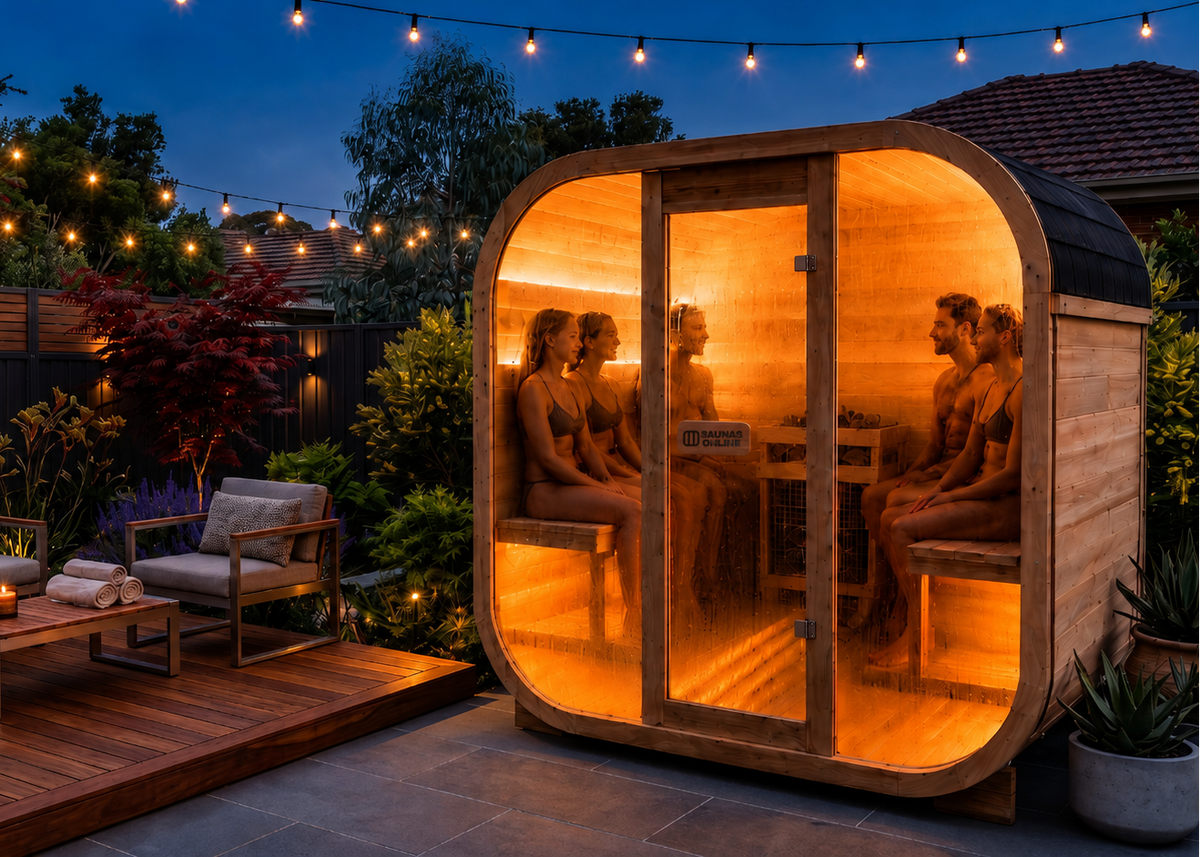 A round wooden outdoor sauna cabin with glowing warm interior lights, shown beside patio seating at dusk. Several people are visible inside the sauna, and the scene includes a deck, string lights, plants, and a dark evening sky.